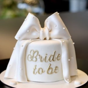 Minimalist white bridal shower cake featuring a large white fondant bow, scattered pearls, and gold "Bride to be" text. Custom elegant cake delivery in Hyderabad, Banjara Hills, and Jubilee Hills.
